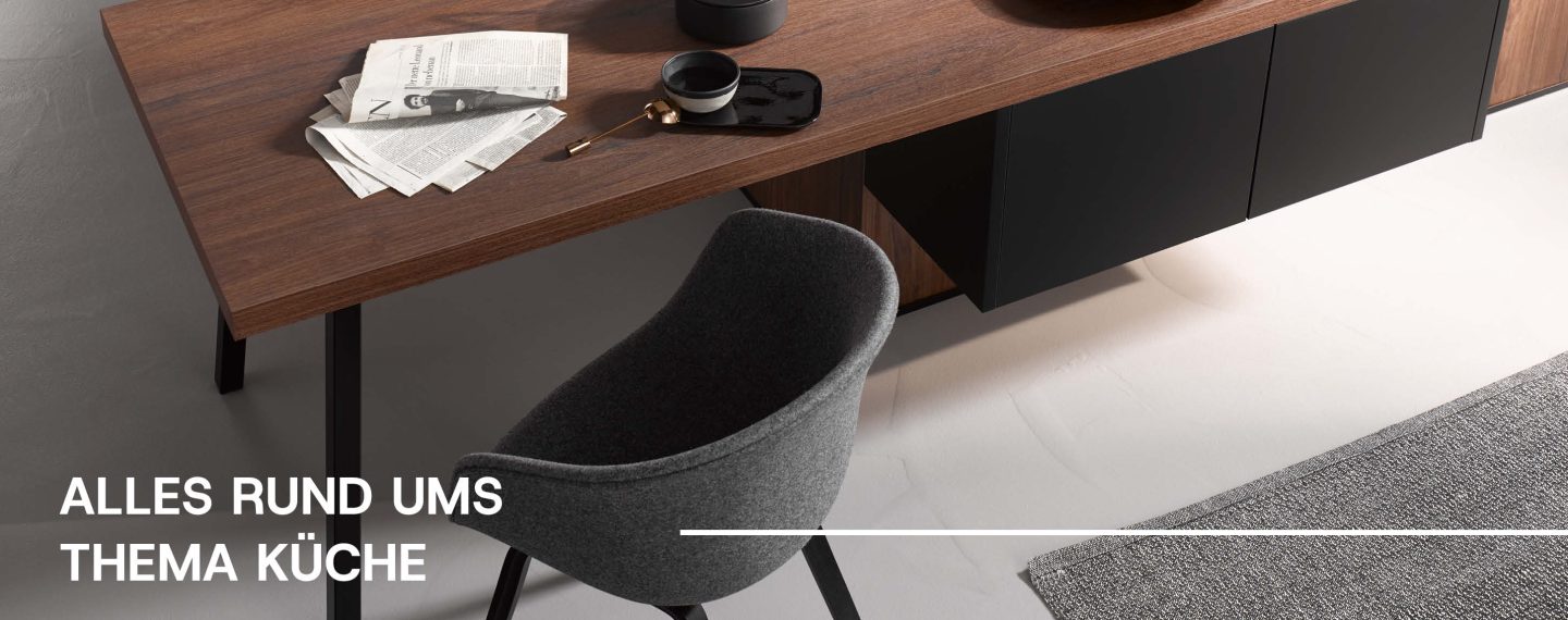 Moderner Stuhl an einem Tisch in Nussbaumoptik. Darauf liegt eine Zeitung und eune Tasse Tee. Darunter steht 