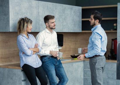 Drei Leute unterhalten sich in einer grauen L-Küche mit Holzarbeitsplatte. – Küche kaufen in Langwedel