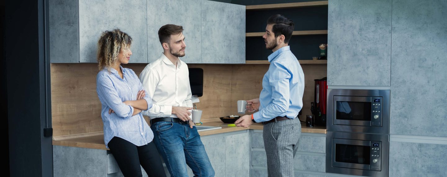 Drei Leute unterhalten sich in einer grauen L-Küche mit Holzarbeitsplatte. – Küche kaufen in Langwedel