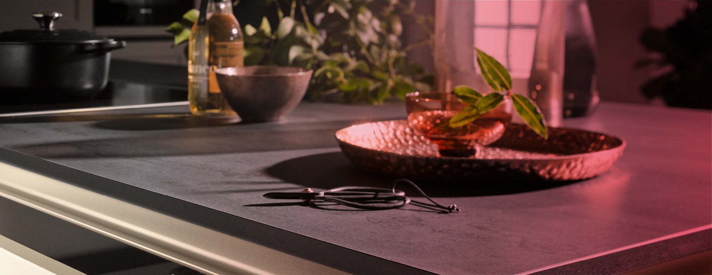 Nahaufnahme einer dunklen Arbeitsplatte mit einem Bronzefarbenen Teller und einem Cocktail darauf. Darüber liegt ein roter Farblayer.