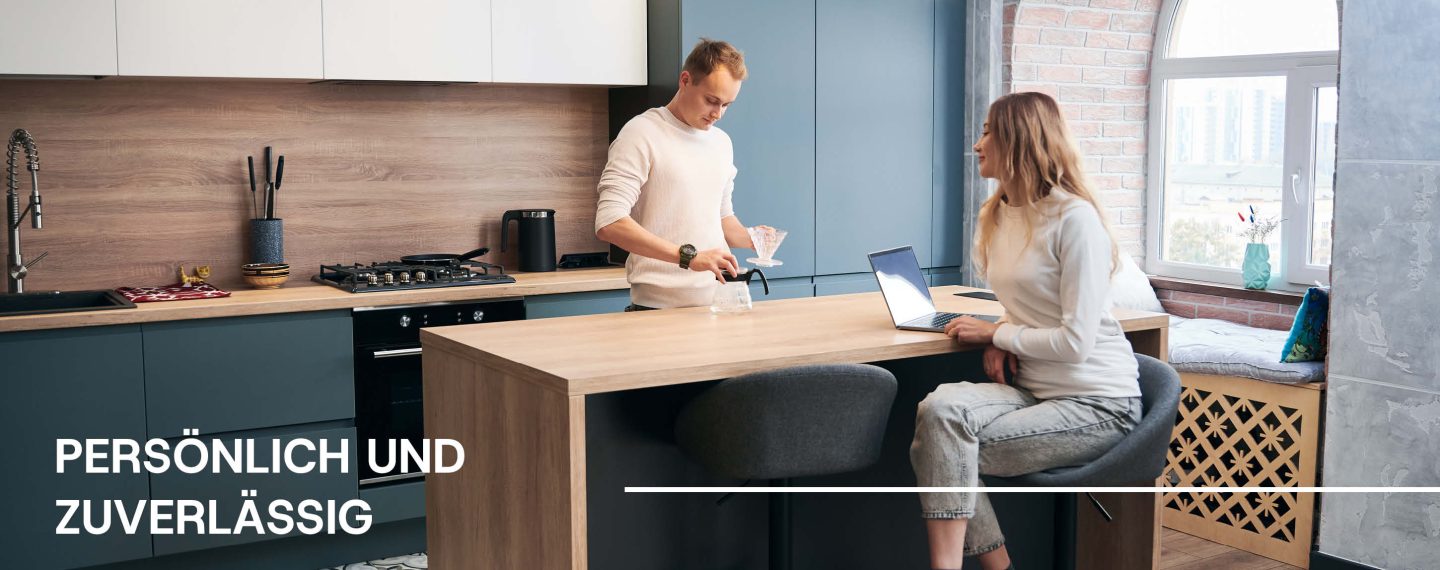 Pärchen in einer blauen, grifflosen Küche mit Kücheninsel. Er befüllt eine Kaffekanne, sie arbeitet an einem Laptop. Daruaf steht 