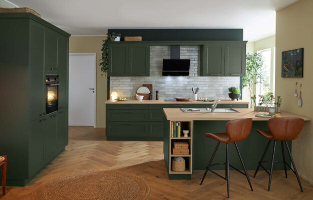 Landhausküche von Schüller in mattem Grün mit Holzarbeitsplatte, Kücheninsel mit Sitzplätzen, Einbaugeräten und klassischer Kassettenfront im warmen Wohnambiente