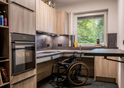 Moderne barrierefreie Küche mit Rollstuhl, unterfahrbarer Arbeitsplatte, höhenverstellbarem Tisch, ergonomisch angeordnetem Kochfeld und Backofen sowie hellen Küchenmöbeln in Holzoptik.