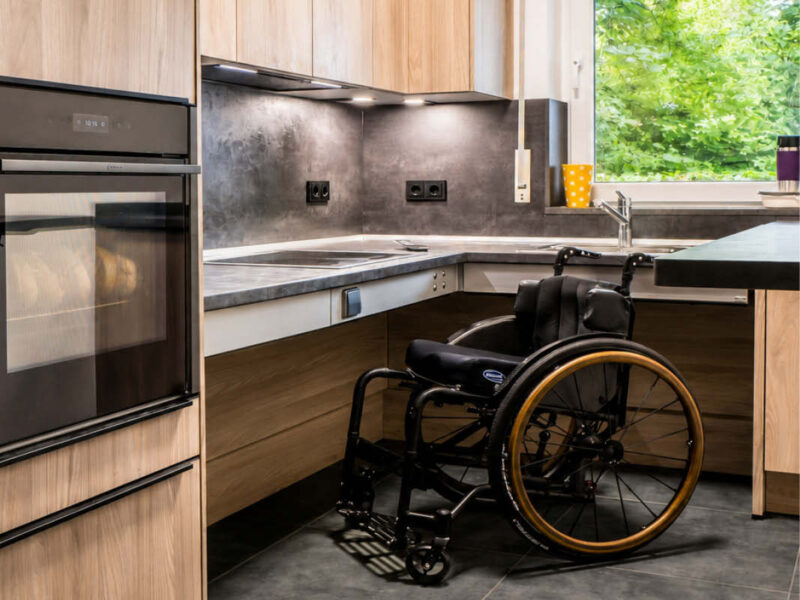 Moderne barrierefreie Küche mit Rollstuhl, unterfahrbarer Arbeitsplatte, höhenverstellbarem Tisch, ergonomisch angeordnetem Kochfeld und Backofen sowie hellen Küchenmöbeln in Holzoptik.